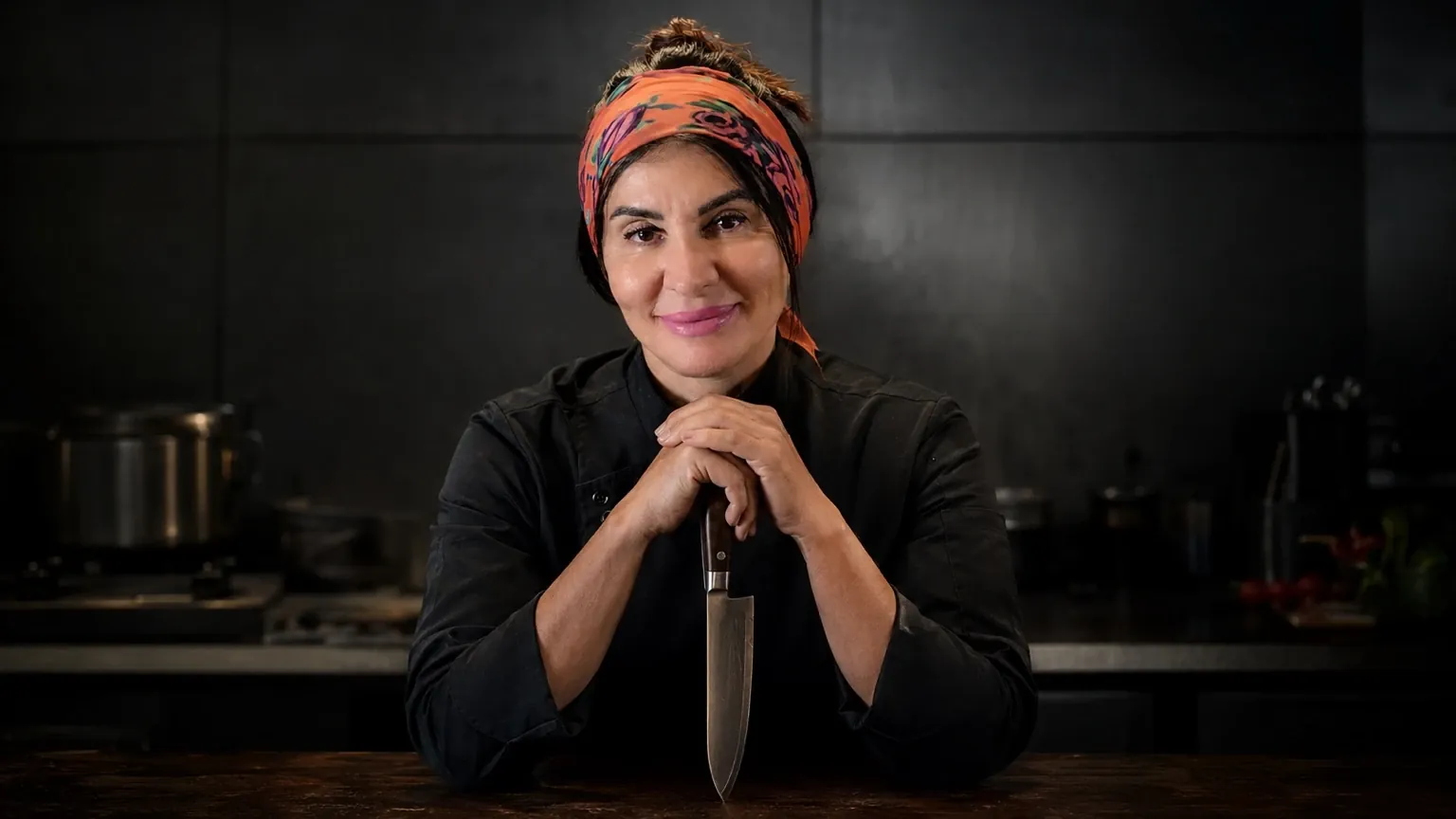 Retrato da Chef Regina Maatouk sorrindo, com lenço laranja e faca de cozinha apoiada na mesa
